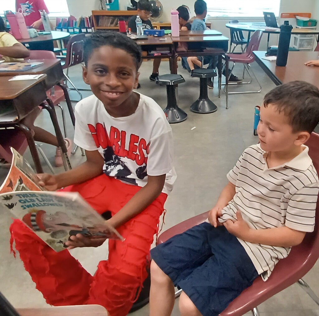 Two children in a classroom, one reading a book while the other listens, seated in red chairs.