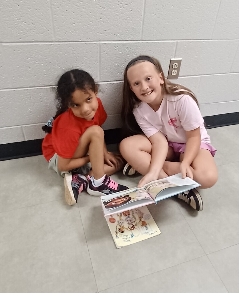 Two girls sit on the floor reading a book, one holding it, both wearing casual clothing.