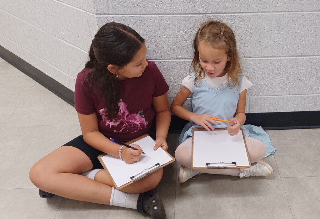 Two young girls sit on a floor, one with a pen, the other holding a clipboard. They are in a hallway with a white wall.