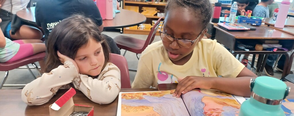 Two children at a table. One reading a book, the other with their head resting on their arm.