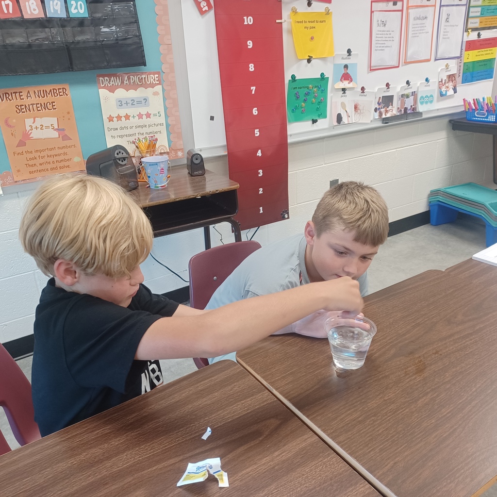 Two young boys conduct a science experiment with a clear liquid in a classroom. A bulletin board and classroom bulletin board are behind them.