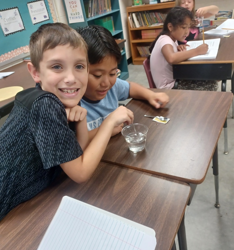 Two boys sitting at a table with a glass of water. Another child sits behind them at another table.