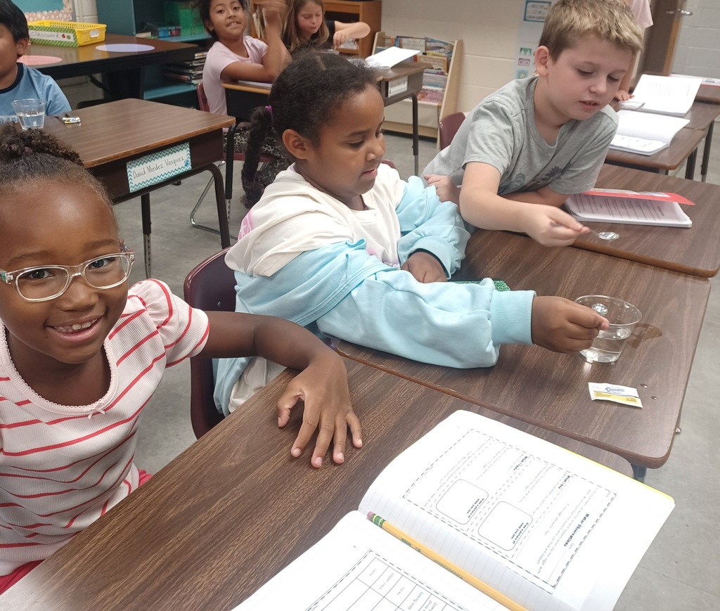 Several children sit at desks in a classroom, with one girl in glasses smiling. A boy leans over to look at something in a cup.