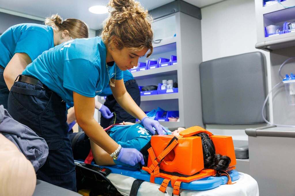 EMT students practice patient immobilization and movement techniques, working together to safely secure a simulated patient to a stretcher during hands-on training.