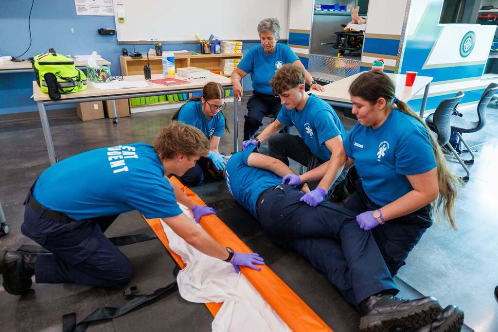 EMT students practice safely rolling and positioning a simulated patient while preparing patient-moving equipment during hands-on training.