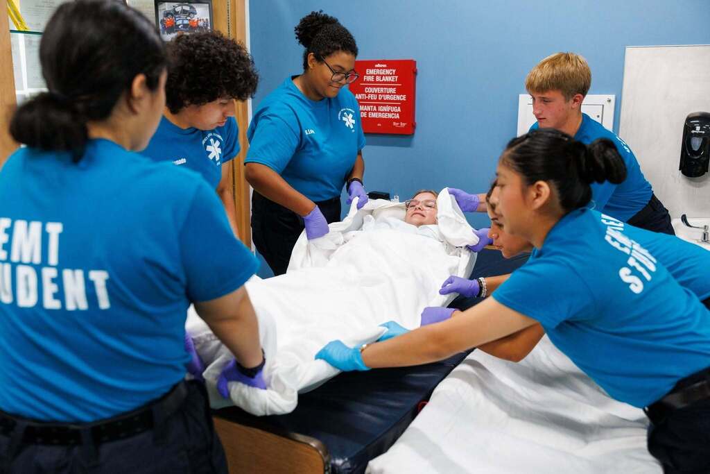EMT students work as a team to safely move a simulated patient from a bed while practicing proper patient lifting and transfer techniques.