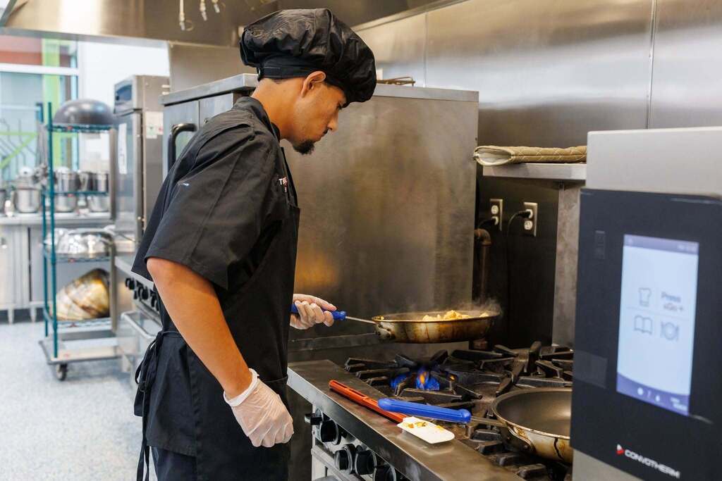A Culinary Arts student develops professional cooking skills at the range, practicing sautéing techniques and gaining hands-on experience with commercial kitchen equipment.