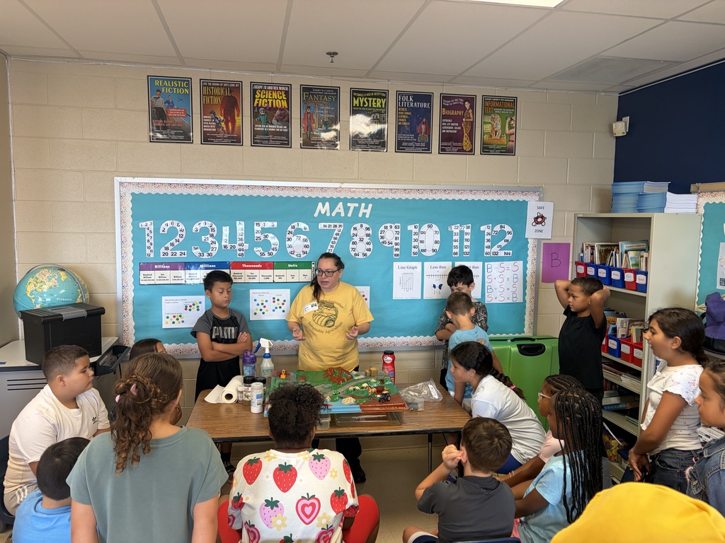 A classroom with a math bulletin board. A teacher with a yellow shirt stands in front of a table. Children sit around the table and listen.