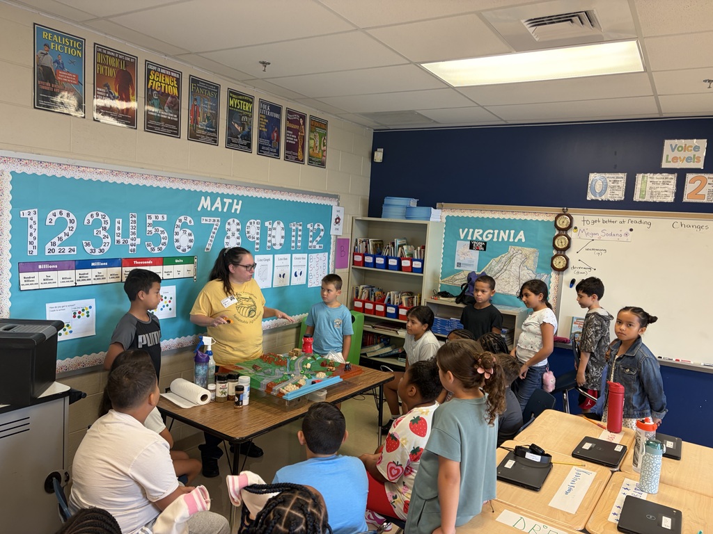 A classroom with a teacher in a yellow shirt, standing by a table with children sitting around it.