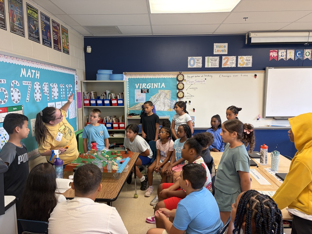 A classroom with students seated in rows, a teacher standing near a table, and a whiteboard with colorful charts.
