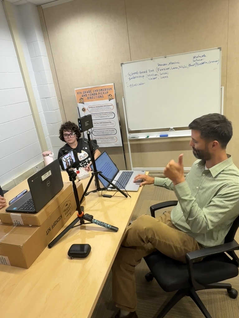 People sit at a desk with laptops, a whiteboard, and boxes. One person gestures.