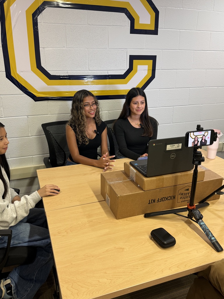 Three individuals sit at a desk, one using a laptop. Two are facing a camera mounted on a tripod. A wall behind them features a large letter C.
