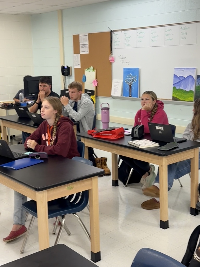A classroom with students seated at desks, using laptops. A white board is on the wall with papers and art.