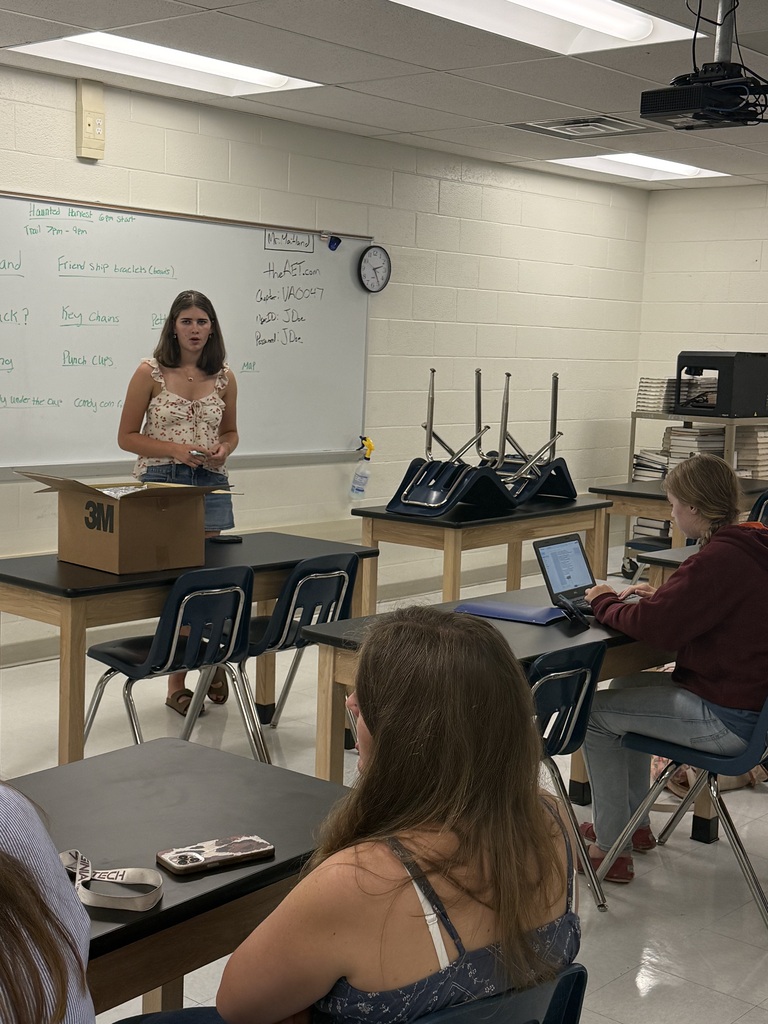 A classroom with a woman standing in front of a whiteboard. Students sit at desks. A projector and clock are on the wall.