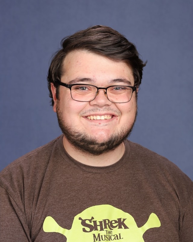 A man with glasses and a beard wears a brown shirt with a yellow Shrek graphic.