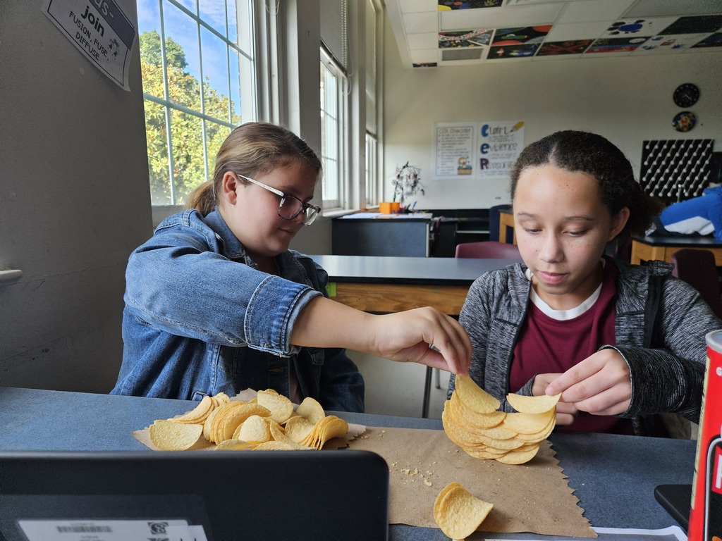 Two young women work with potato chips on a table in a classroom with large windows.