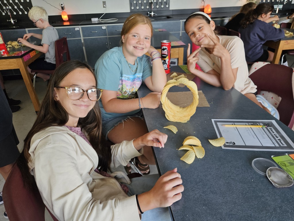 Students seated at a table, one holding a yellow object, another posing for the camera. Table has papers and plastic containers.