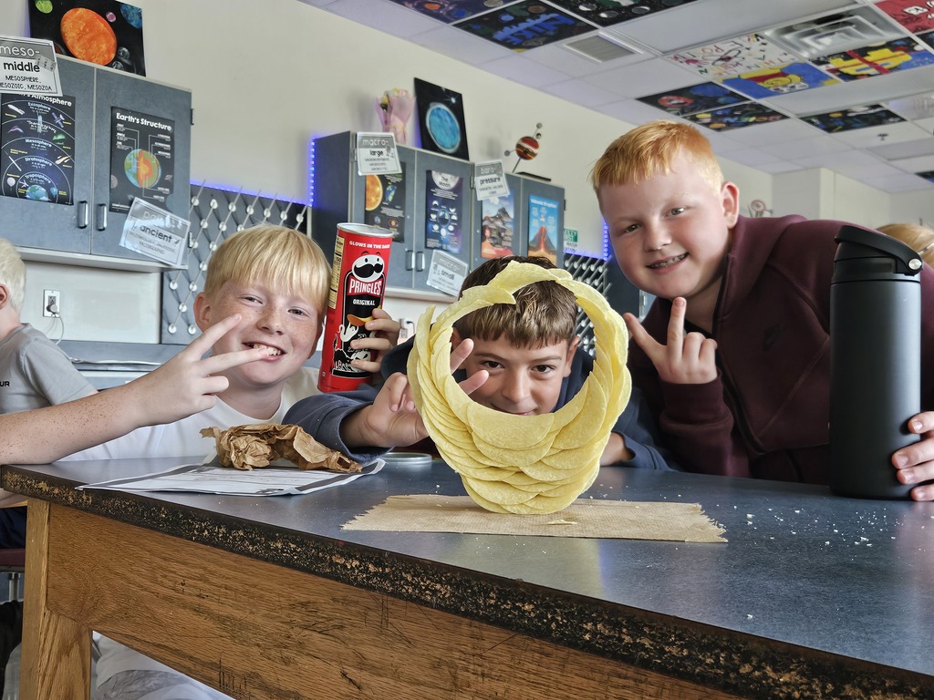 Three boys pose behind a table with one holding a yellow object. One boy wears a red shirt.