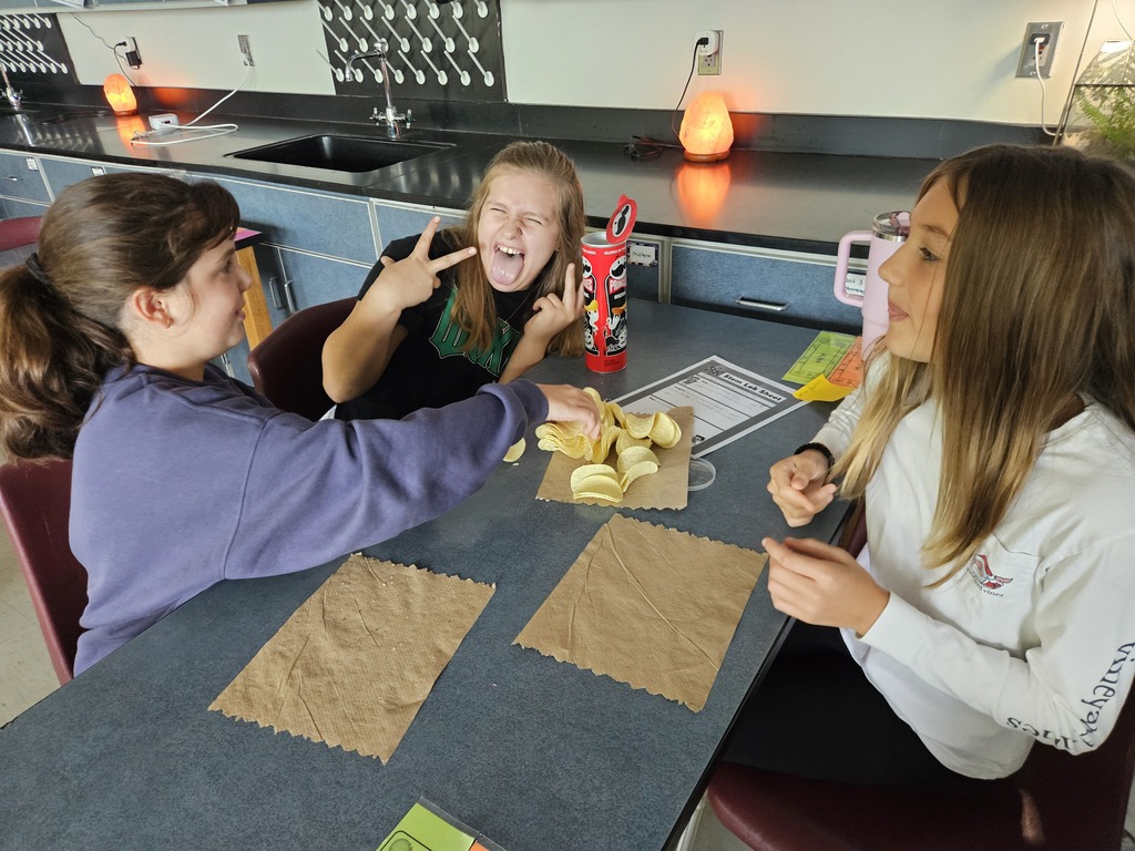 Three people sit at a table with food. One person eats, another makes a peace sign, and the third looks at the first.
