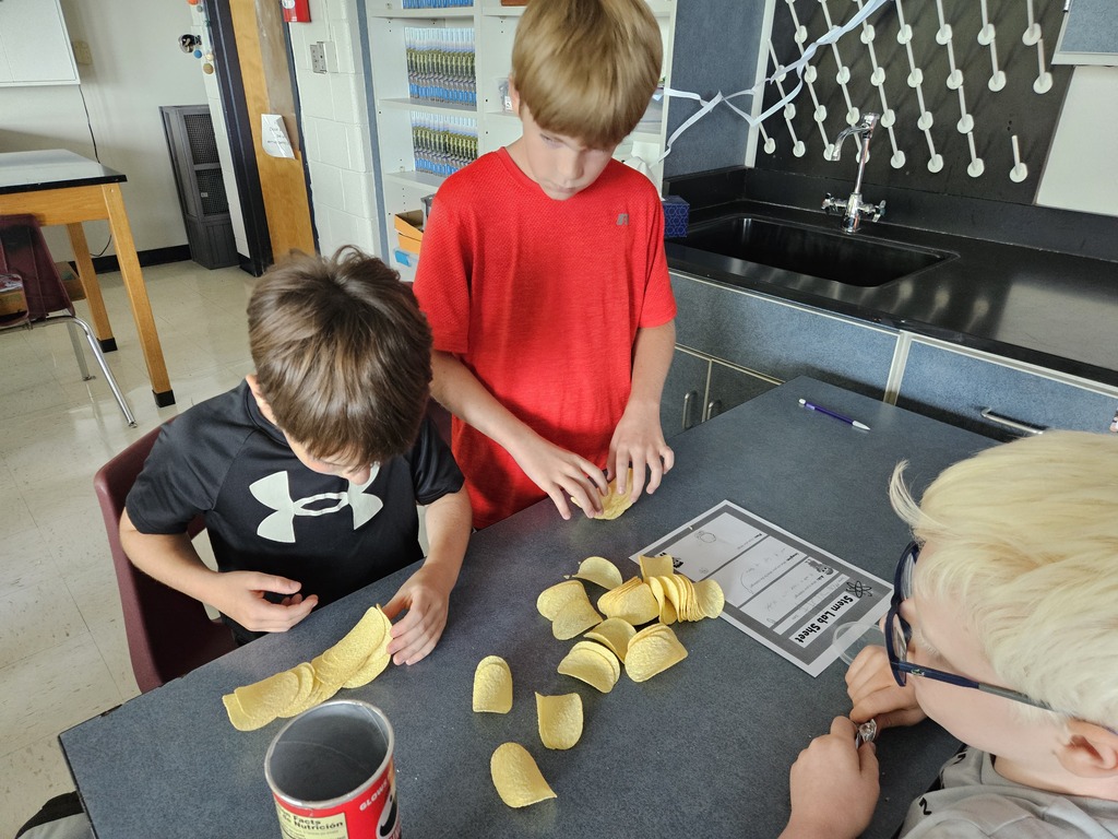 Three children are at a table in a classroom, one making pasta. The table has pasta and a cup.