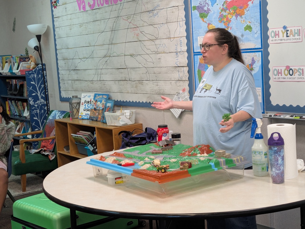 Woman in glasses explaining a model to the class. A table with a model on it and a bookshelf in the background.
