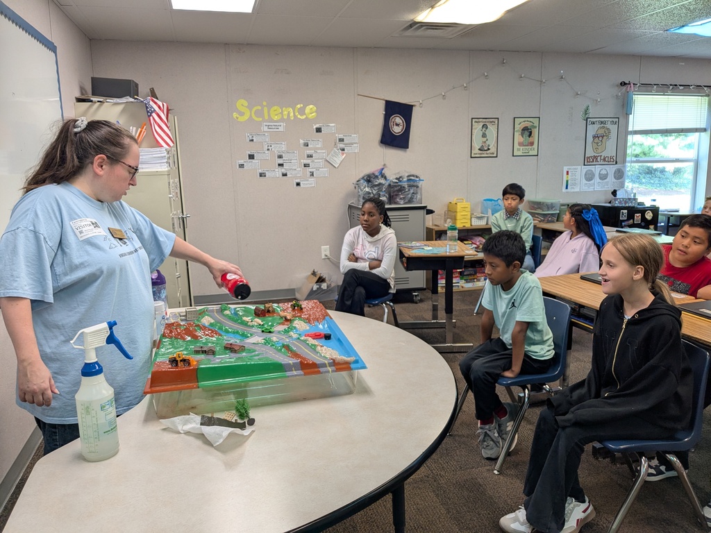 A science teacher sprays water onto a model on a table while a group of children observe.