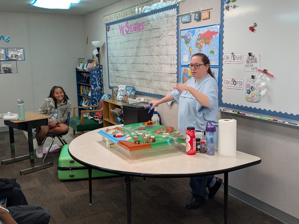A woman stands at a table in a classroom, pointing to a model while students sit in chairs.