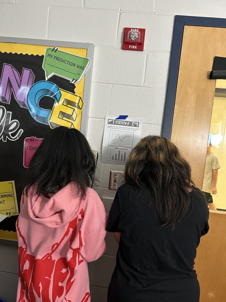 Two people in a room with a bulletin board. One wears a pink hoodie, the other a black shirt.