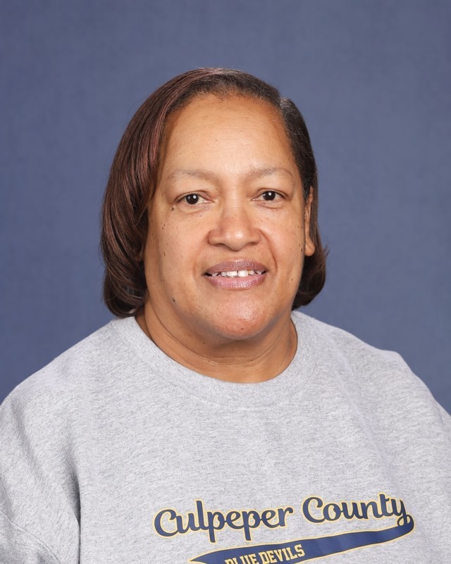A woman with short brown hair smiles at the camera. She wears a gray sweatshirt with "Culpeper County" and "Blue Devils" on it.