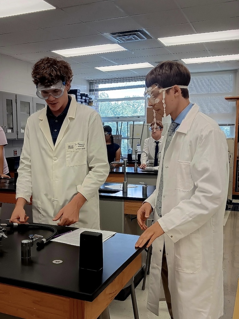 Two individuals in white lab coats and safety goggles work at a table in a science lab.