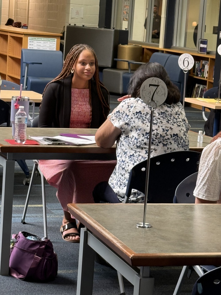 People seated at a table in a room with bookshelves in the background. One woman looks at another woman.