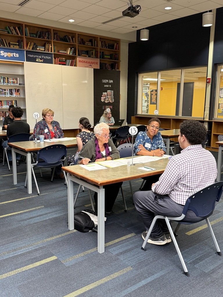 A group of people in a library setting, seated at tables, with some wearing name tags.
