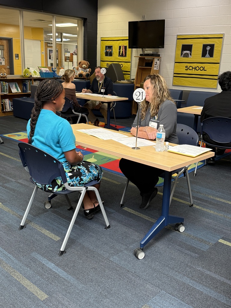 Two women in a classroom setting. One woman is seated while the other stands, both are looking at papers.
