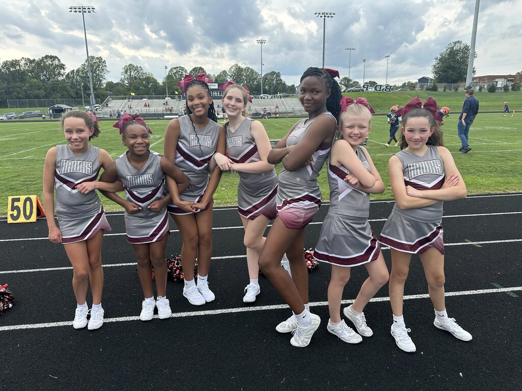 Seven cheerleaders in matching uniforms pose on a track. Behind them, a grassy field and bleachers.
