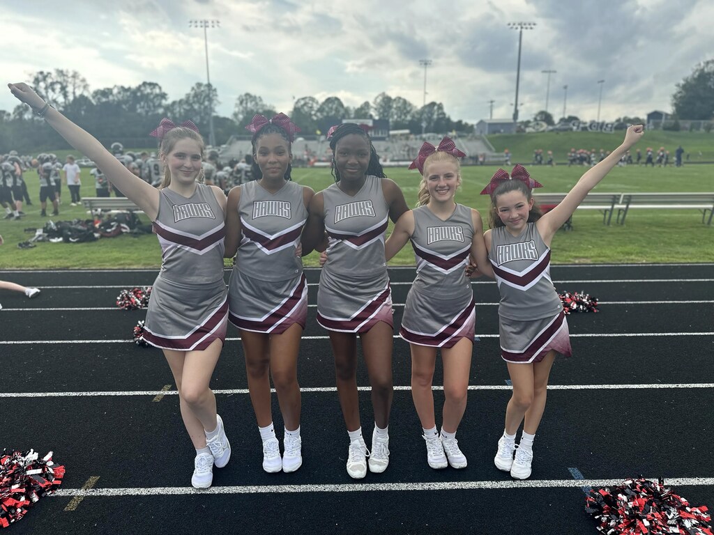 Five young girls in cheerleading uniforms with arms raised on a track with pom-poms in the background.