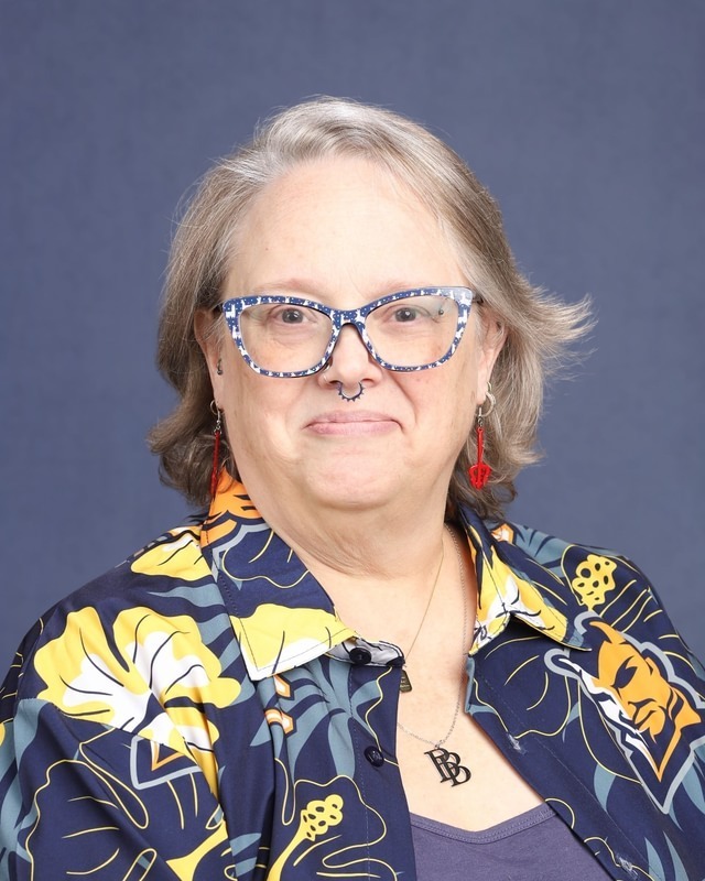 A woman with gray hair wearing glasses and a floral button-up shirt poses against a blue background.