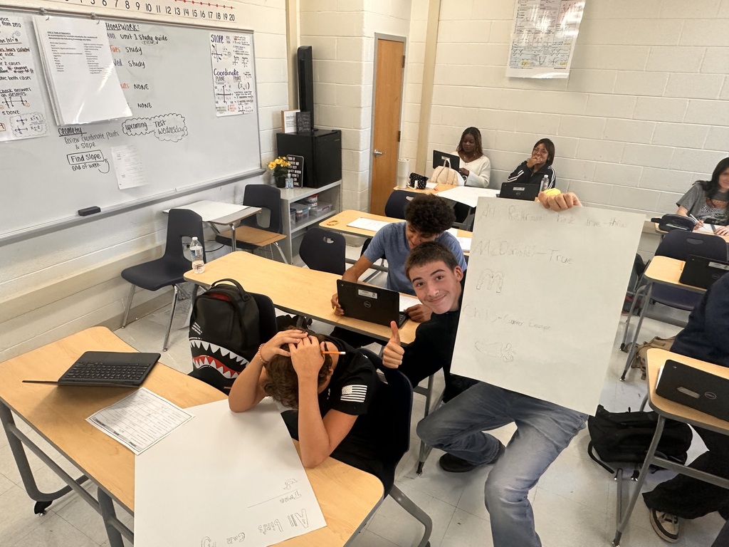 A classroom with students seated. Two students are holding up a piece of paper. There is a whiteboard on the wall.