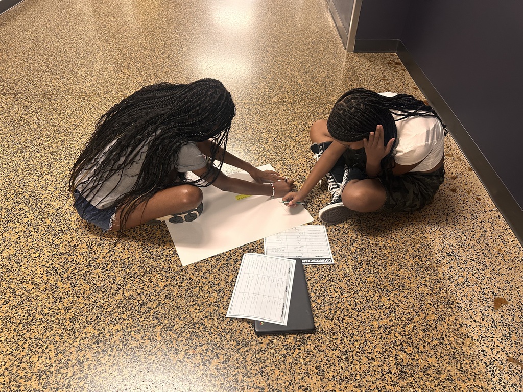 Two people kneel on a speckled floor, looking at a large piece of paper with notes and a tablet.