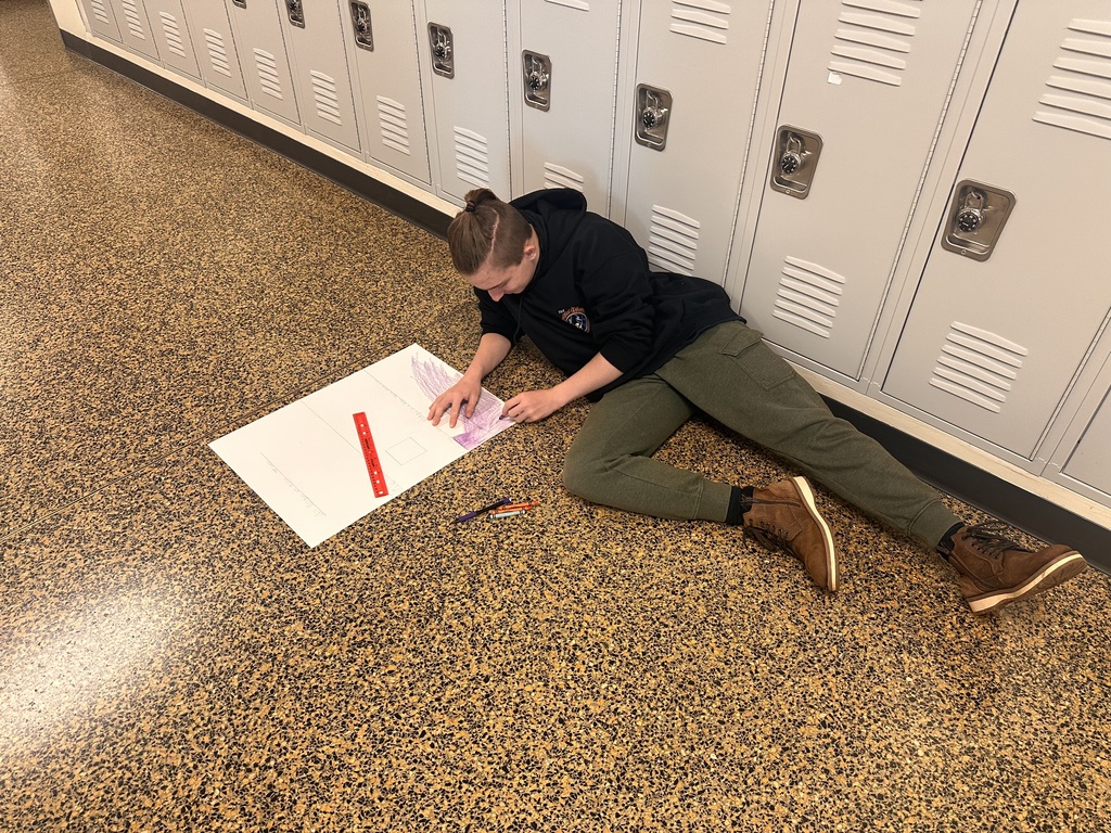 Person lying on the floor next to lockers, holding a paper and colored pencils.