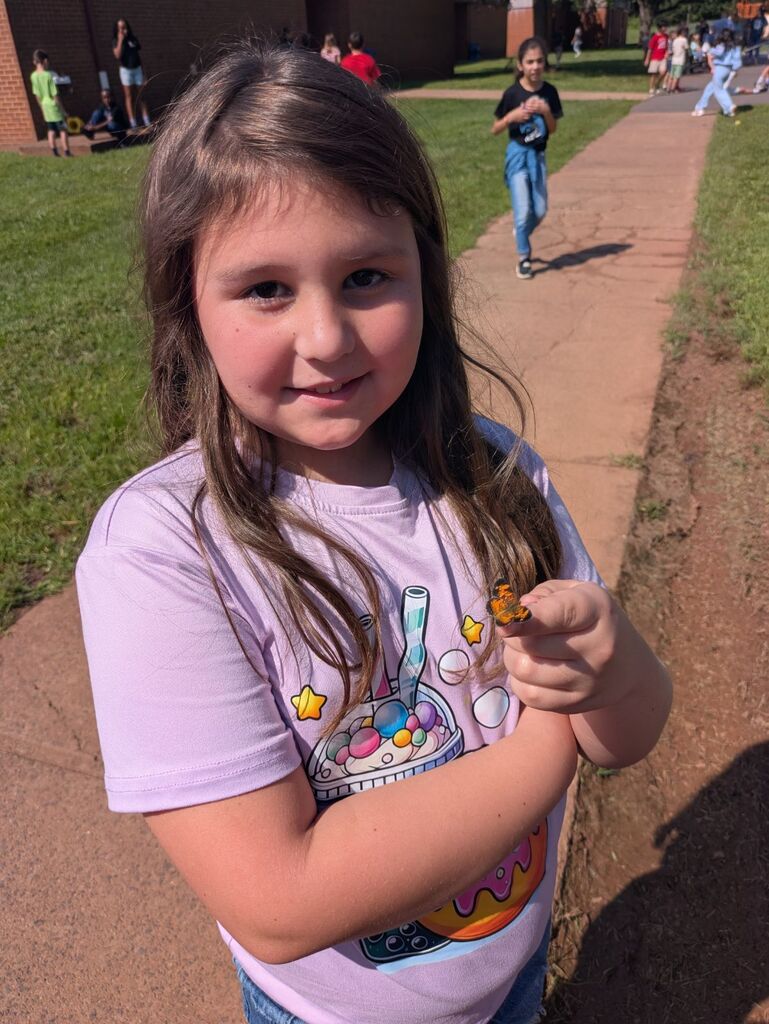 A young girl in a purple shirt with a cake print holds a small object. She stands on a sidewalk, with grassy areas and people in the background.