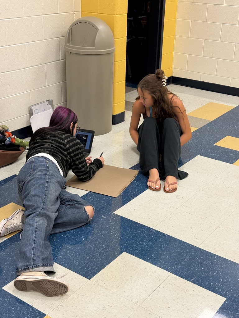 Two women sit on a floor, one with a tablet, the other taking notes. A trash bin is nearby.
