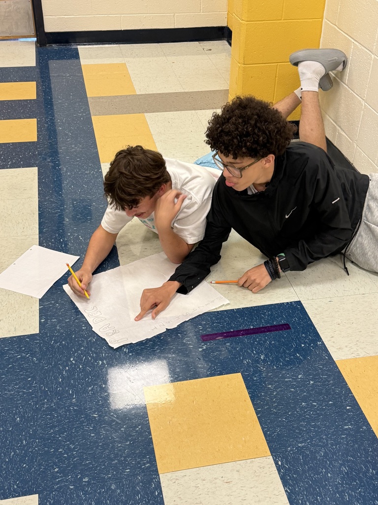 Two young people lie on a floor, using paper and pencils to draw on the floor.
