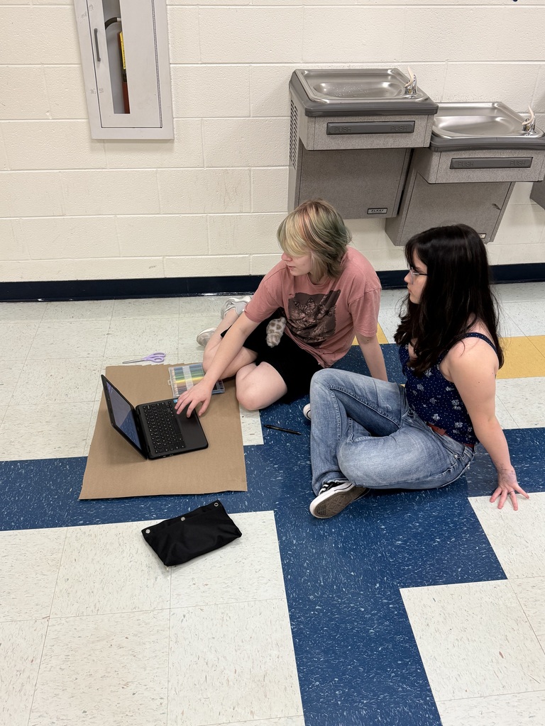 Two people sitting on the floor with a laptop, a bag, and a wall with sinks in the background.