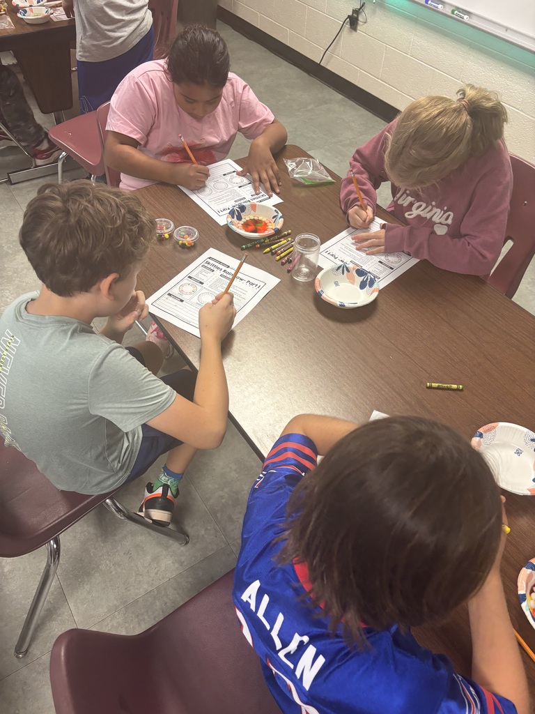 Four children sit around a table in a classroom, drawing on paper. A white plate and markers are on the table.