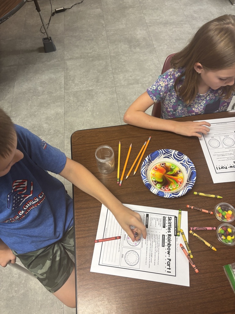 Two children work at a table with papers, crayons, a bowl of candy, and a cup.