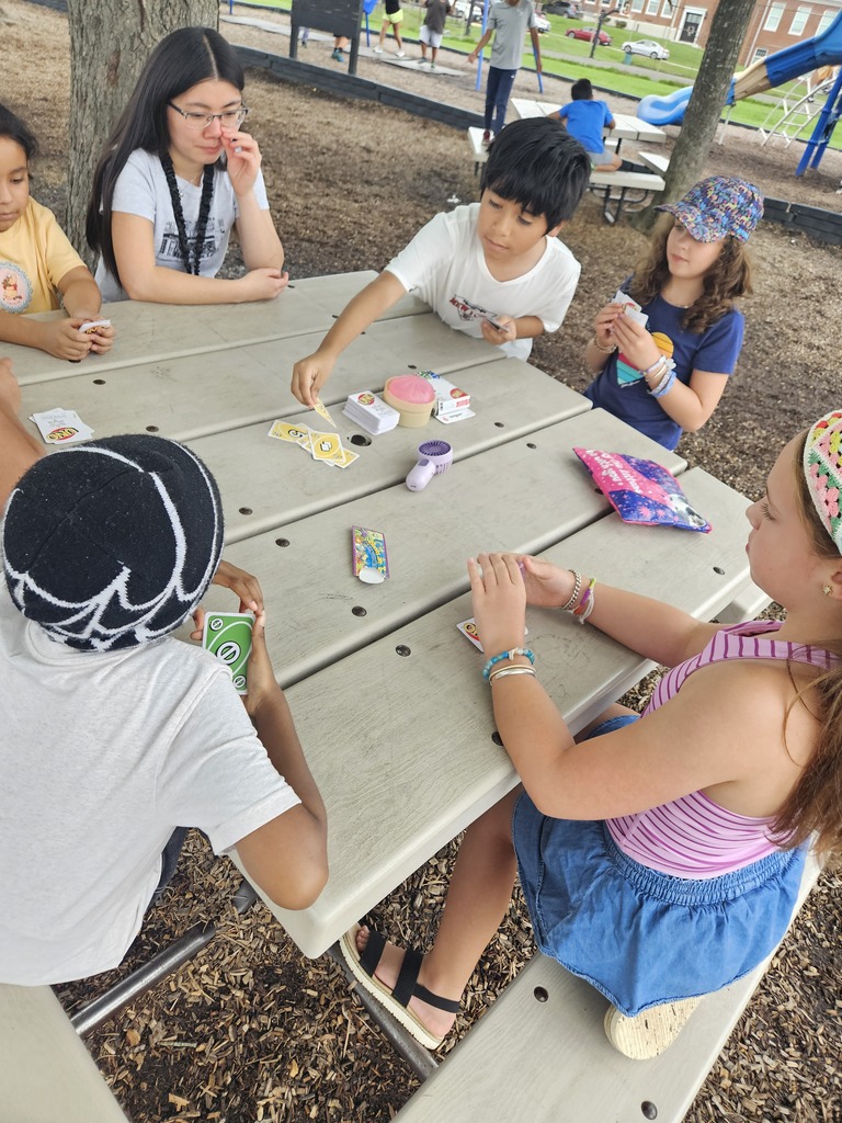 Children seated around a picnic table in a park, playing a card game. Some wear bracelets and glasses.