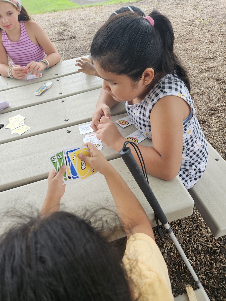 A few girls sit around a picnic table. Two of them hold cards and look at them.