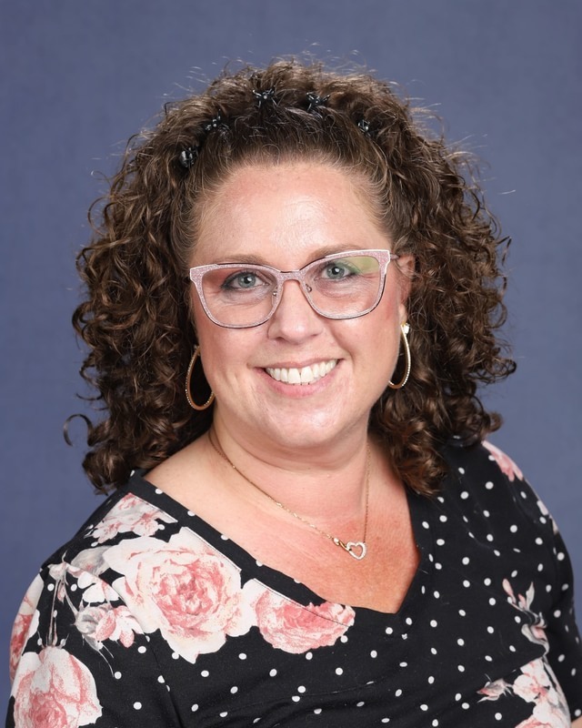 A woman with curly hair wearing glasses, a floral polka-dot shirt, and a necklace. She smiles at the camera.