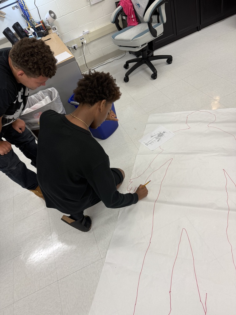 Two individuals work on a large white sheet with red lines, likely in a classroom setting.