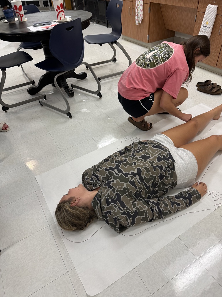 A female traces another as she lies on paper on the floor of a classroom.